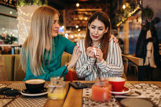 Upset young woman crying at a cafe table while her attentive friend provides comfort and emotional support during a difficult time, holding her shoulder and offering a tissue