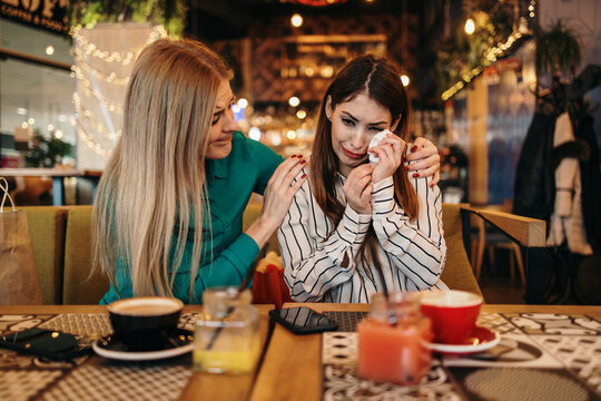 Upset young woman crying at a cafe table while her attentive friend provides comfort and emotional support during a difficult time, holding her shoulder and offering a tissue