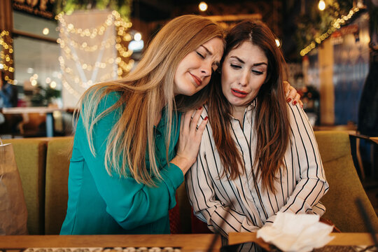 Upset young woman crying at a cafe table while her attentive friend provides comfort and emotional support during a difficult time, holding her shoulder and offering a tissue