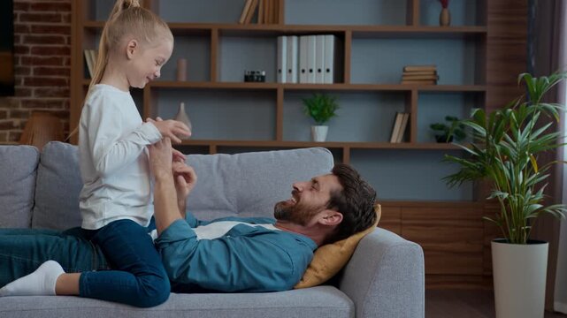 Caucasian family at home loving father with little playful girl tickling dad with active child kid daughter playing tickle on couch sofa funny entertainment in living room enjoying spending good time