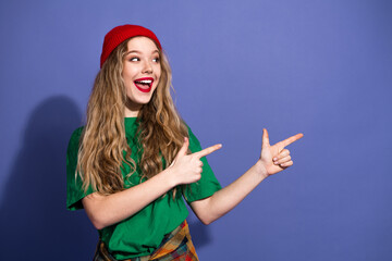Happy young woman with red beanie and green shirt points playfully at something off camera against a purple backdrop
