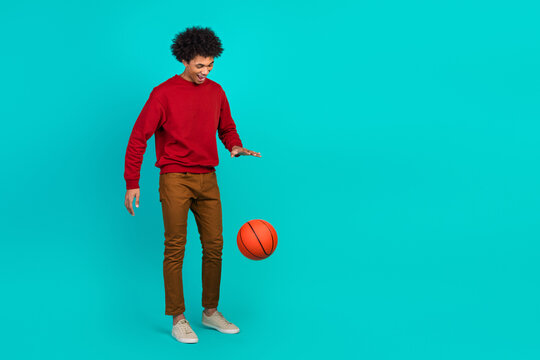 A young man in a red sweater plays with a basketball in a bright turquoise studio smiling with joy and casual style