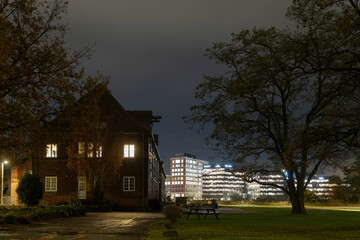 Naklejka premium An illuminated historic brick building and a large tree frame a grassy park contrasting with brightly lit modern office towers under a dark night sky.