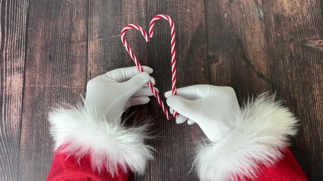 Santa's gloved hands holding two striped candy canes against a wooden background, joining them to form a heart, conveying a festive spirit