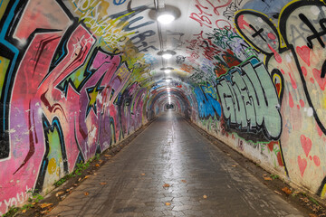 A long, arched pedestrian tunnel is illuminated by overhead lights and features vibrant, colorful street art across its walls and ceiling.