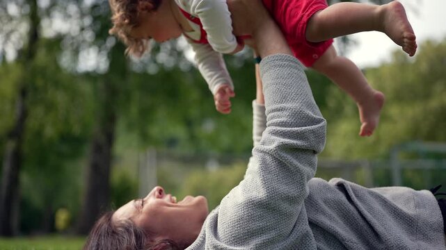 Mother lying on grass lifting baby joyfully in air, both smiling and laughing together in sunny park, expressing love, warmth, and playful connection between parent and child