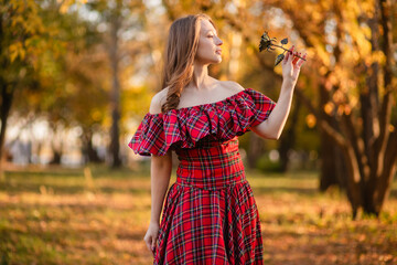 A young woman stands in an autumn forest. She is wearing a long, red, plaid dress with off-the-shoulder details and a frill, a romantic, vintage style. She holds a decorative metal flower in her hands