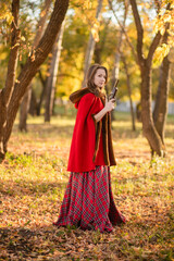 A young woman stands in an autumn forest. She's wearing a long red plaid dress and a warm cape, a romantic, vintage style. She's holding a pistol.