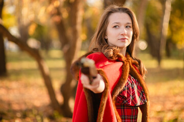 A young woman stands in an autumn forest. She's wearing a long red plaid dress and a warm cape, a romantic, vintage style. She's holding a pistol.