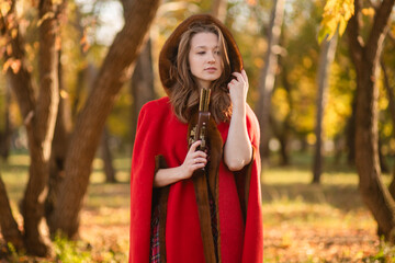 A young woman stands in an autumn forest. She's wearing a long red plaid dress and a warm cape, a romantic, vintage style. She's holding a pistol.