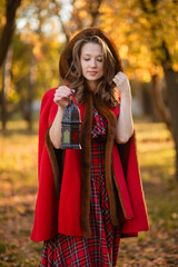 A young woman stands in an autumn forest. She's wearing a long red plaid dress and a warm cape, a romantic, vintage style. She's holding a lantern.