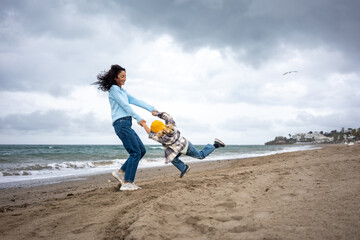 Joyful mother swinging her young son by the arms on the sandy beach, playful family moment by the sea with happy child in yellow beanie and woman in blue sweater