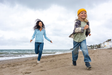 Smiling mother and cheerful young son running barefoot along the sandy beach, enjoying playful family time together by the sea under cloudy sky with ocean waves in the background