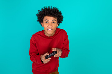 Young man with curly hair in a red sweater plays a game controller against teal background and...