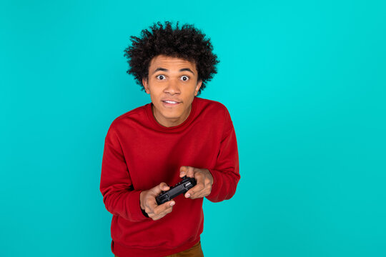 Young man with curly hair in a red sweater plays a game controller against teal background and looks surprised