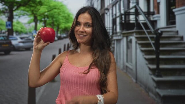 Woman smiling while holding red apple on a city street near building steps and parked cars; healthy happiness.
