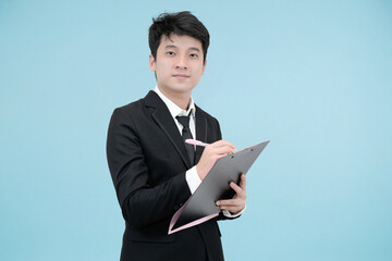 A cheerful and diligent young Asian businessman in a black suit, smiling while writing or checking a document on a clipboard against a light blue studio background