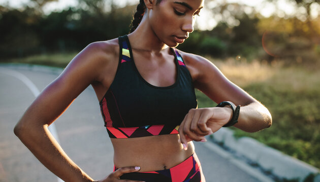 Focused young woman in athletic wear checking her fitness tracker on her wrist during an outdoor workout, monitoring her progress under the sun