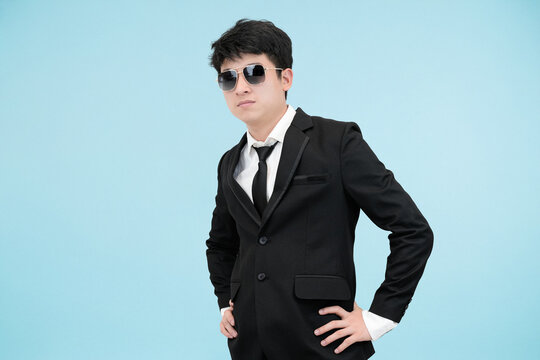 A confident and serious young Asian man in a sharp black suit and sunglasses, standing with his hands on his hips, projecting a cool pose in light studio background