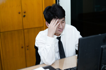 A stressed Asian office worker or businessman sitting at his desk, burying his face in his hands,...