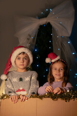 Children in festive sweaters with Santa hats looking from a big gift box surrounded by Christmas lights and tinsel