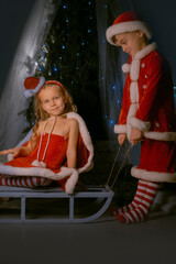 Brother and sister enjoying the holiday season as the boy rides the girl on a sled by a sparkling Christmas tree indoors.