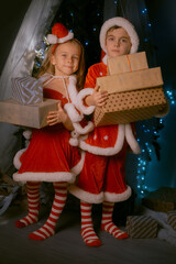 Cute siblings in festive elf costumes standing by a Christmas tree with a big stack of wrapped gifts.