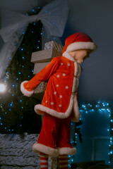 Child in Santa costume and hat near a festive tree with blue lights, carrying beautifully wrapped presents behind him