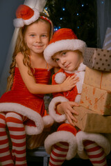 Cute siblings in festive elf costumes enjoying the holiday season while sitting on a sled filled with Christmas gifts by a sparkling tree.