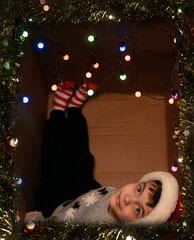 Boy in Christmas sweater lying upside down in box with twinkling lights having fun and enjoying festive holiday mood
