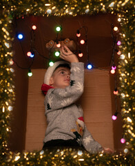 Boy playing with a wooden toy car on top of a decorated gift box with lights and tinsel, festive holiday mood
