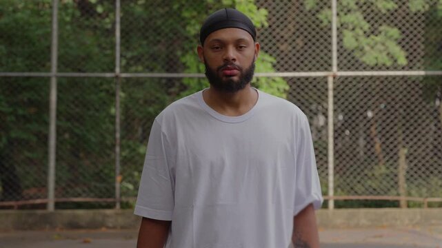 African American man wearing durag standing outdoors in basketball court, looking directly at camera with serious calm expression and composed presence against wire fence