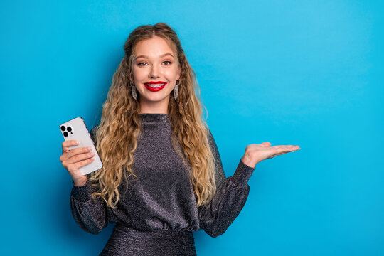Glamorous young woman wearing a shiny dress holds a smartphone on a bright blue background for a stylish fashion and tech lifestyle shot - Powered by Adobe