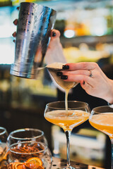 Close-up of a bartender pouring an amber cocktail through a strainer from a metal shaker into elegant coupe glasses on a bar counter, selective focus
