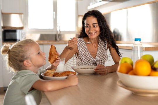 Smiling mother enjoying cereal breakfast while looking at her child eating croissant, happy family morning routine with milk, fresh pastries and fruits on the kitchen table
