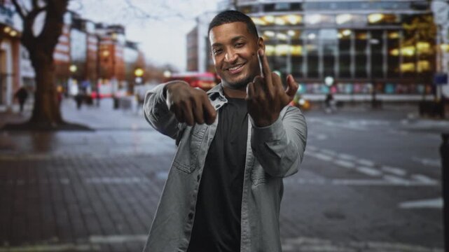 Young black man shows middle finger and points with hand on wet city street by building, smiling; defiant confidence.