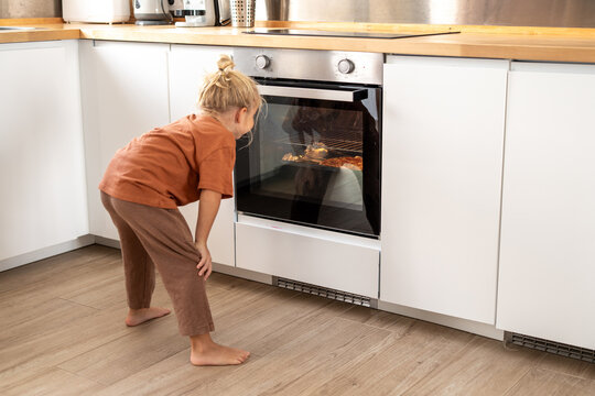 Curious young boy in brown clothes bending forward and looking into oven while waiting for food to bake in modern bright kitchen - Powered by Adobe