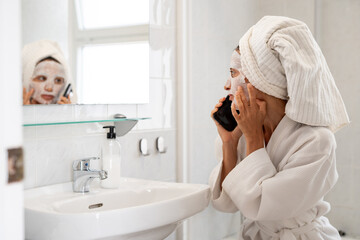 Woman in bathrobe with sheet mask on face talking on smartphone in bathroom near sink and mirror