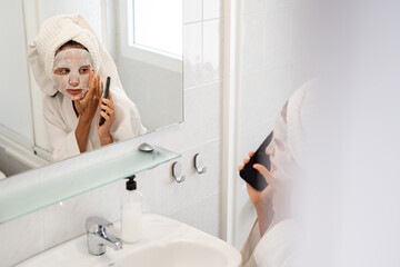 Woman in bathrobe with sheet mask on face talking on smartphone in bathroom near sink and mirror
