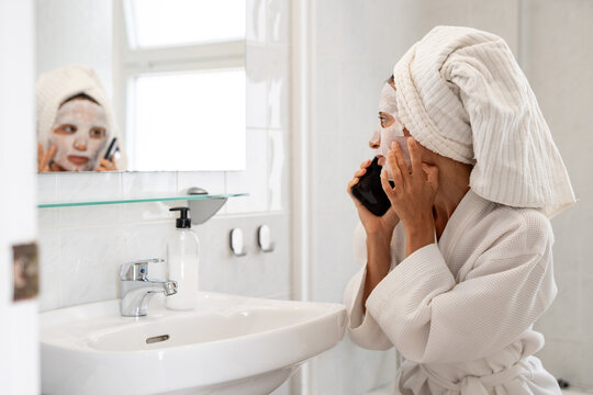 Woman in bathrobe with sheet mask on face talking on smartphone in bathroom near sink and mirror