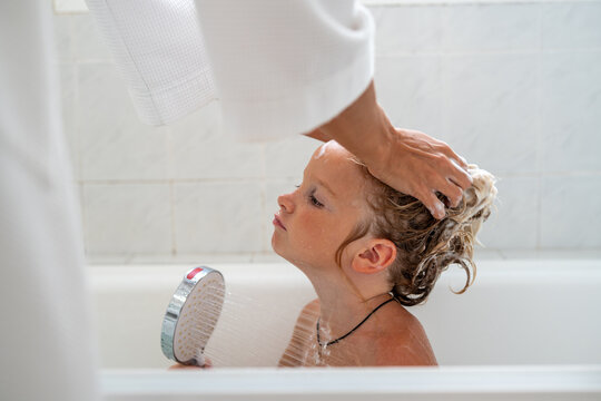 Little boy in bathtub holding shower head while mother washing his hair with shampoo foam and water running down his face