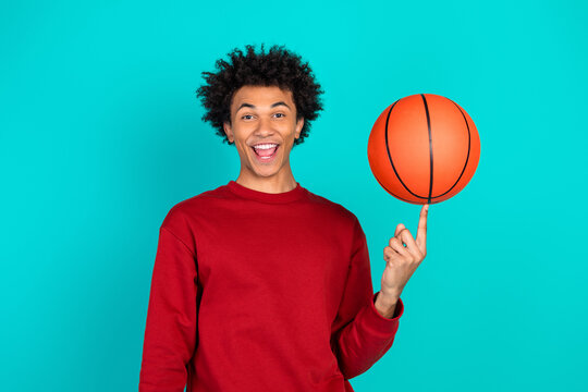 Happy young man spins a basketball on his finger against a teal background wearing a red sweater and smiling - Powered by Adobe