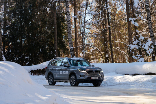 Issyk-Kul, Kyrgyzstan - December 13, 2024: A gray MG RX8 SUV with illuminated DRLs and promotional branding drives on a snow-covered road flanked by tall trees on a sunny winter day