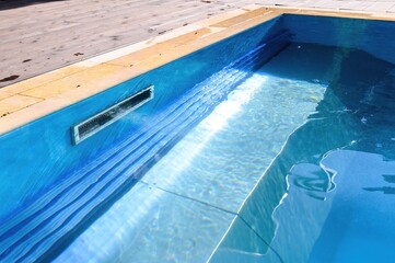 A skimmer installed in the pool wall removes debris from the water surface, maintaining cleanliness. Bright sunlight creates shimmering reflections on the blue water