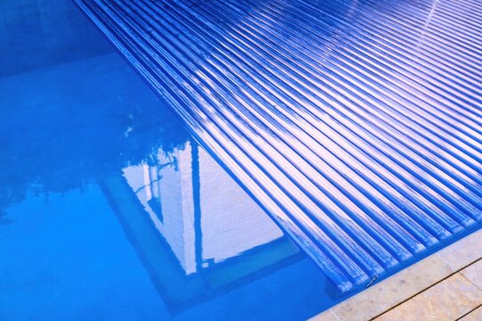 A swimming pool partially covered with a blue slatted cover. The reflection of a house on the water surface creates an interesting visual effect. Morning or evening light in a private backyard