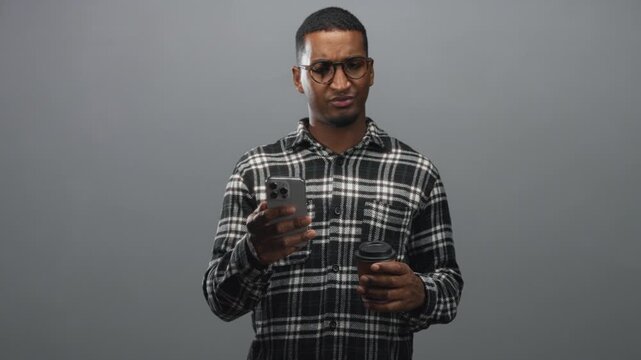 Black man wearing glasses and plaid shirt holding a smartphone in one hand and a takeaway coffee cup in the other, frowning while reading the screen in studio; confusion.