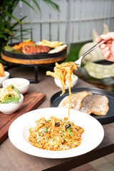 A hand lifting chicken spaghetti coated in rich tomato sauce with a fork, captured with softly blurred seafood dishes in the background, creating a warm and appetizing dining-scene presentation.