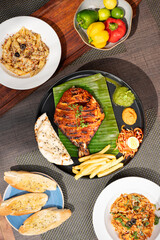 Top shot of tandoori pomfret masala served with chutney, salad, fries, and kulcha on a plate, styled alongside chicken spaghetti, cheesy penne seafood, garlic bread, and raw capsicums 