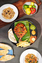 Top shot of tandoori pomfret masala served with chutney, salad, fries, and kulcha on a plate, styled alongside chicken spaghetti, cheesy penne seafood, garlic bread, and raw capsicums 