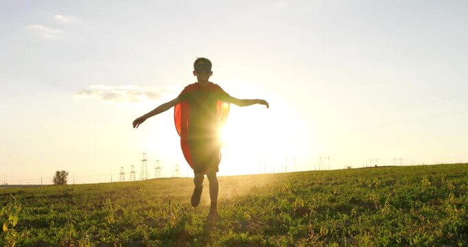 Child boy runs in red raincoat into field at sunset, confidently and boldly spreading his arms. Symbol of heroism life goals. Childhood readiness to help others and dream of becoming strong and kind